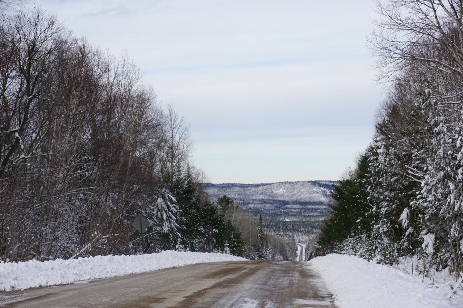 A snow-banked two-lane road running downhill between winter forest towards low forested hills